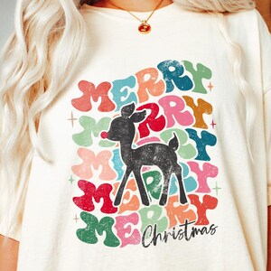 May include: A white t-shirt with a colorful graphic design of the word "Merry" repeated in a circle with a reindeer in the center. The text "Christmas" is below the circle.