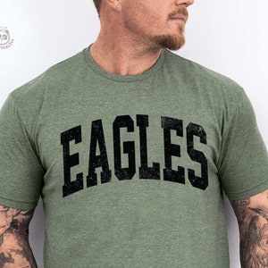 K&ouml;nnte beinhalten: Olivgr&uuml;nes T-Shirt mit dem Wort "EAGLES" in fetter, schwarzer, verwaschener Schrift. Das Shirt hat einen Rundhalsausschnitt und scheint aus einem weichen, melierten Stoff zu sein. Das Shirt wird von einer Person getragen.