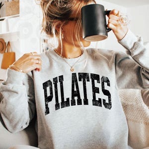 May include: A light gray Pilates sweatshirt with the word "PILATES" in bold, black, distressed lettering. The sweatshirt is paired with matching gray sweatpants. A black mug is held up to the face. The overall theme is fitness and activewear.