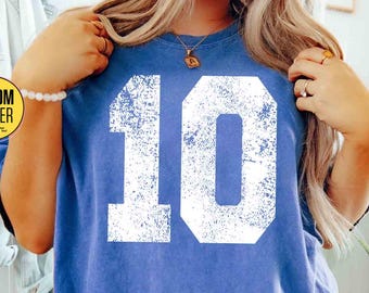 May include: A blue t-shirt with the number "10" in white, distressed font. The shirt has short sleeves and a crew neck. The text "CUSTOM NUMBER" is in a yellow circle. The color is "FLO BLUE".
