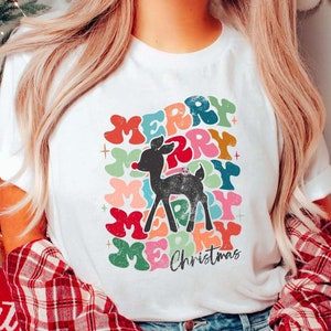 May include: White Christmas t-shirt featuring a retro-style reindeer graphic. The design incorporates multicolored 'Merry Christmas' text in a layered, overlapping arrangement. The reindeer is black, and the text is in various shades of red, green, yellow, orange, pink, and blue. The overall style is vintage and festive, suitable for the Christmas season.