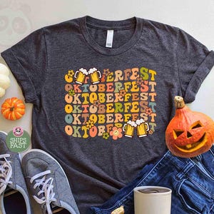 May include: A dark grey t-shirt with the word "Oktoberfest" in a retro font, decorated with beer mugs and flowers. The shirt is styled with blue jeans, grey trainers, and carved pumpkins, creating an autumnal theme.
