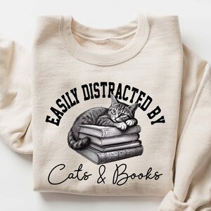 May include: A beige sweatshirt with the text "Easily Distracted By Cats & Books" in black. A black and white illustration of a cat sleeping on a stack of books is above the text.