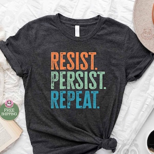 Puede incluir: Una camiseta gris oscuro jaspeada con el texto "RESIST. PERSIST. REPEAT." en una fuente de estilo vintage. El texto está en naranja, verde y azul.