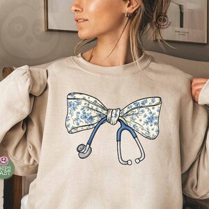 Op de afbeelding: Beige sweatshirt met een blauw en wit bloemenstrikontwerp en een stethoscoop. De strik is het centrale beeld, met de blauwe slang en zilveren oortelefoons van de stethoscoop. De sweatshirt is een casual, comfortabele stijl.