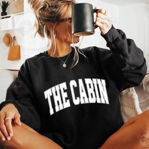May include: A black sweatshirt with white text that reads "THE CABIN" in a collegiate font. The sweatshirt is being worn by a person sitting on a couch.