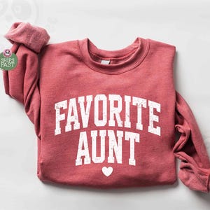 以下が含まれることがあります： 白いブロック体で「FAVORITE AUNT」と書かれた、小さな白いハートが付いた赤みがかったスウェットシャツ。長袖トップスは白い表面にきれいに折りたたまれています。ブランドロゴは左下に表示されています。