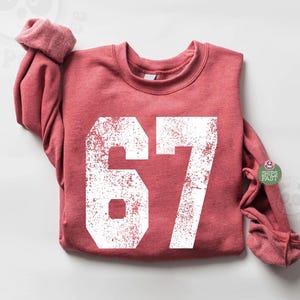 Puede incluir: Una sudadera de color rojo anaranjado con el número 67 en una fuente blanca desgastada. La sudadera tiene mangas largas y cuello redondo. El texto "Ships Fast" está en una pequeña pegatina en la manga.