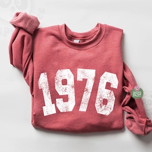 May include: A red sweatshirt with the year "1976" printed in large, distressed white letters. The long-sleeved top has a crew neck and a relaxed fit. The image includes a "Ships Fast" sticker.