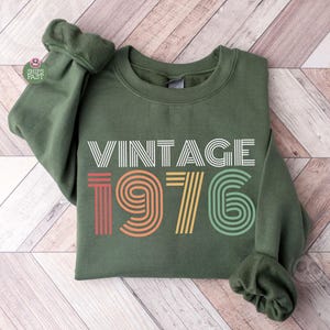 May include: An olive green sweatshirt featuring the text "VINTAGE 1976" in white, red, orange, yellow, and green. The sweatshirt is folded on a light wood-grain surface. The sleeves are bunched up.
