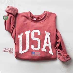 May include: Heather scarlet red sweatshirt with the word "USA" in distressed white lettering and a small American flag graphic. The sweatshirt has a crew neck and long sleeves.