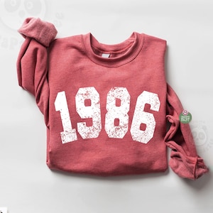 May include: A red sweatshirt with the year "1986" printed in large, distressed white letters. The long sleeves are rolled up. The sweatshirt is displayed on a white surface. The overall theme is a vintage or retro style.