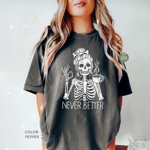 May include: A pepper-colored t-shirt featuring a white graphic of a skeleton with a messy bun hairstyle, holding a coffee cup and making an okay hand gesture. The text "NEVER BETTER" is printed in white below the skeleton illustration.
