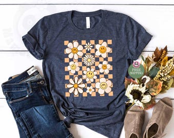 May include: A dark heather navy blue short-sleeve t-shirt featuring a central graphic of a peach and white checkered pattern overlaid with various white daisy flowers, each containing a yellow smiley face in the center, paired with dark blue jeans.