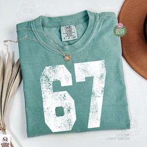 May include: Light green t-shirt with the number 67 in white, featuring a distressed design. The shirt has a crew neck and a tag that reads "COMFORT COLORS". A gold necklace with a pendant rests on the shirt. A brown hat is visible in the background.