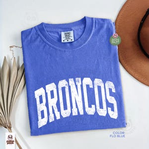 May include: A blue t-shirt with the word "BRONCOS" in large, distressed white letters across the front. The shirt has a crew neck. A brown hat is partially visible in the background. The shirt's colour is described as "Flo Blue".