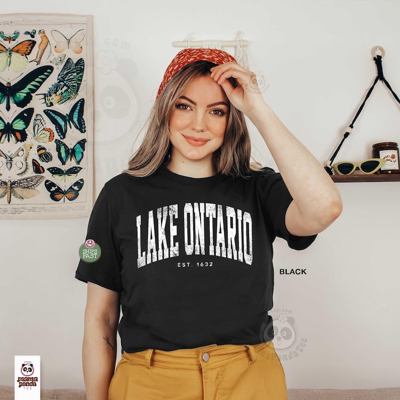 May include: A black short-sleeve t-shirt featuring white distressed text that reads LAKE ONTARIO EST. 1632 across the chest. The shirt is paired with mustard yellow pants. The design has a weathered, vintage-inspired appearance on the fabric.