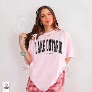 May include: A light pink short-sleeved t-shirt featuring the black text LAKE ONTARIO and EST. 1632 printed in a distressed, arched font across the chest. The shirt has a crew neckline and a loose, relaxed fit, shown in a plain studio setting.