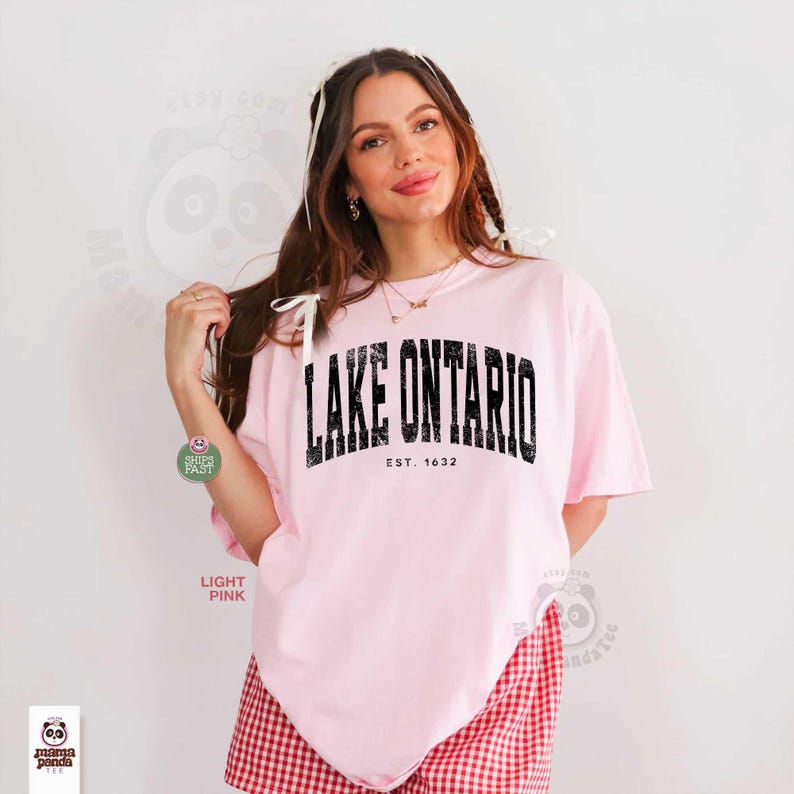 May include: A light pink short-sleeved t-shirt featuring the black text LAKE ONTARIO and EST. 1632 printed in a distressed, arched font across the chest. The shirt has a crew neckline and a loose, relaxed fit, shown in a plain studio setting.