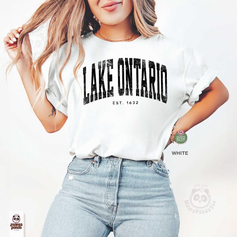 May include: A white short-sleeved t-shirt featuring the black text LAKE ONTARIO and EST. 1632 printed in a distressed collegiate font across the chest. The shirt is paired with light blue denim jeans, creating a casual look with a clear graphic design.