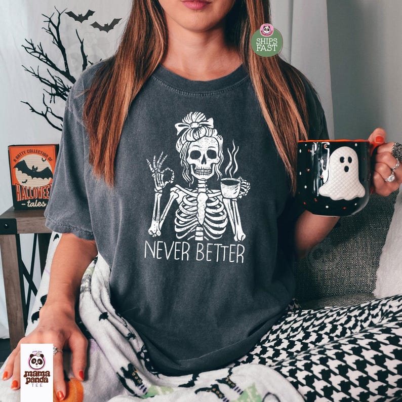 May include: A charcoal gray t-shirt featuring a white graphic of a skeleton with a messy bun holding a coffee cup and making an okay hand gesture. The text below the skeleton reads NEVER BETTER in a white font. The shirt has short sleeves.