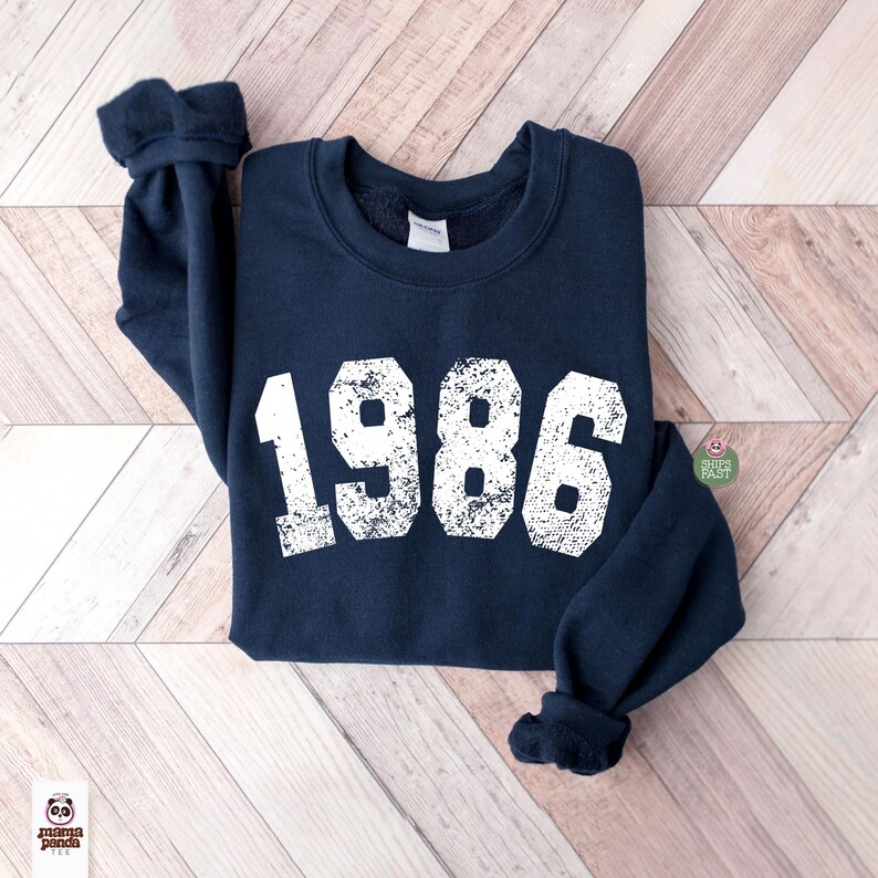 May include: Navy blue sweatshirt with the year "1986" printed in distressed white lettering. The sweatshirt has a crew neck and long sleeves with cuffed wrists. The garment is laid flat on a light wood grain surface.