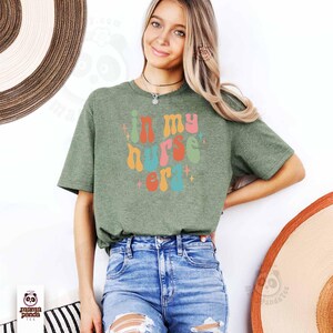 May include: Olive green t-shirt with the text "in my nurse era" in a colorful, retro font. The design is centered on the shirt. The model is wearing blue jeans. The shirt is from Mama Panda Tee.