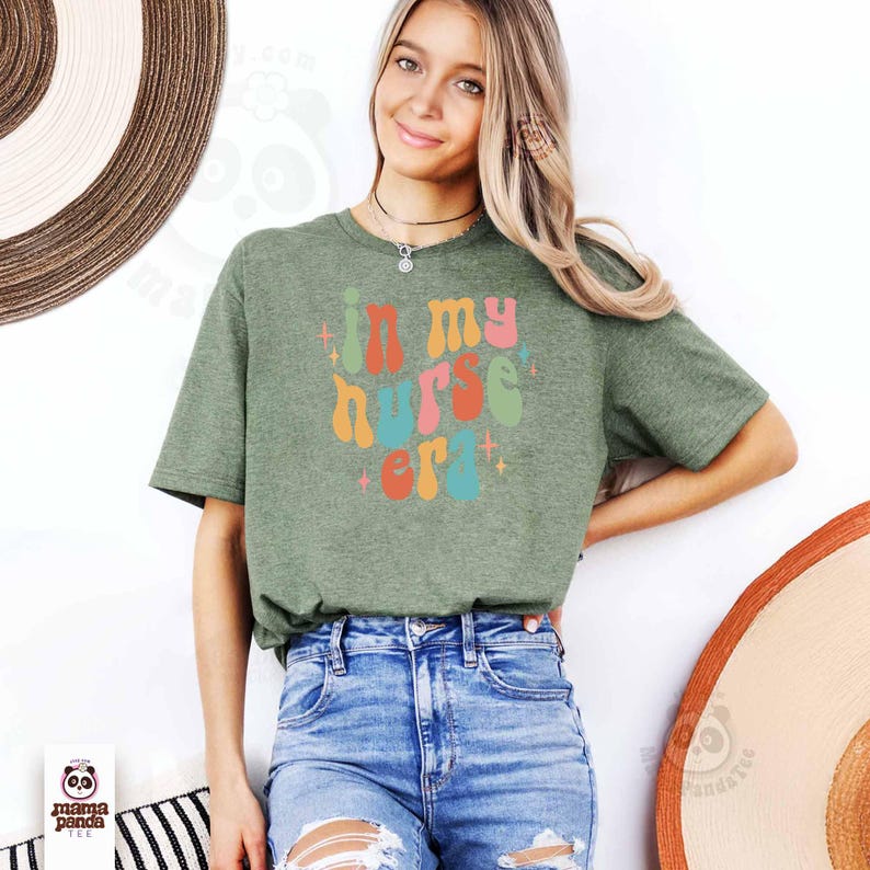 May include: Olive green t-shirt with the text "in my nurse era" in a colorful, retro font. The design is centered on the shirt. The model is wearing blue jeans. The shirt is from Mama Panda Tee.
