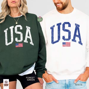 May include: Forest green and white sweatshirts with the word "USA" in distressed white and blue lettering. Each sweatshirt has a small American flag graphic. The forest green sweatshirt is paired with black shorts.