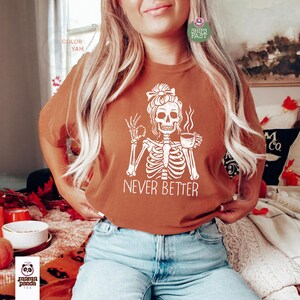 May include: A yam-colored t-shirt featuring a white graphic of a skeleton with a messy bun hairstyle, holding a steaming cup of coffee and making an okay hand gesture, with the text NEVER BETTER printed in white capital letters below the skeleton.