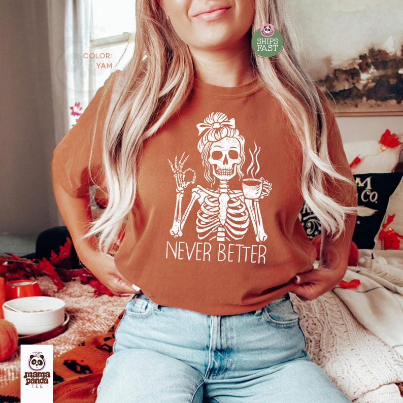 May include: A yam-colored t-shirt featuring a white graphic of a skeleton with a messy bun hairstyle, holding a steaming cup of coffee and making an okay hand gesture, with the text NEVER BETTER printed in white capital letters below the skeleton.