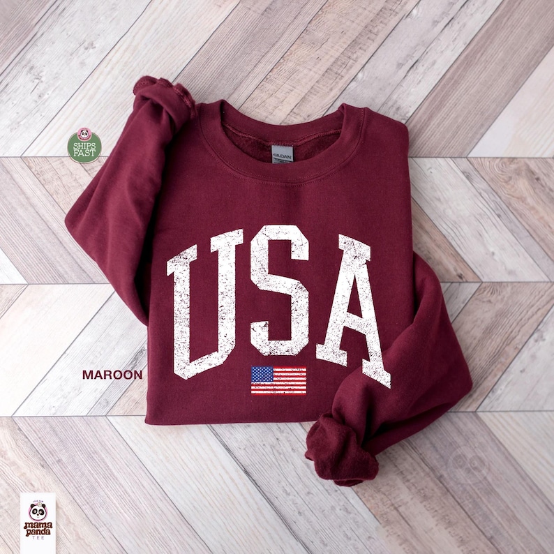 May include: Maroon crewneck sweatshirt with the word "USA" in distressed white lettering and a small American flag graphic. The sweatshirt is folded on a light wood-grain surface.