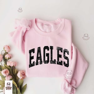 Może przedstawiać: Jasnor&oacute;żowa bluza z nadrukiem "EAGLES" w czarnych, wytartych literach. Bluza jest złożona, widoczne są rękawy. R&oacute;żowe kwiaty ułożone są po lewej stronie zdjęcia.
