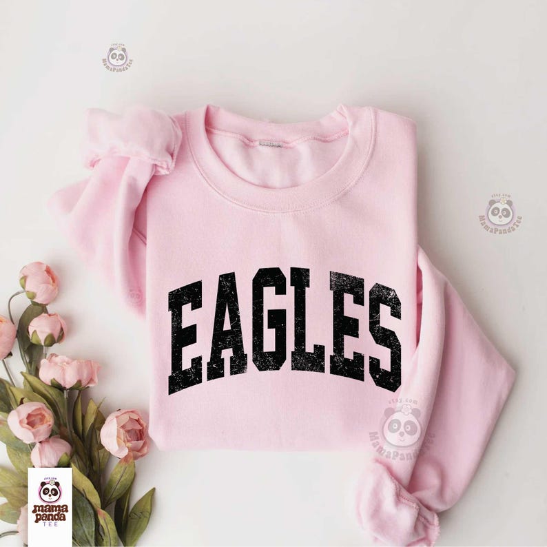 Może przedstawiać: Jasnor&oacute;żowa bluza z nadrukiem "EAGLES" w czarnych, wytartych literach. Bluza jest złożona, widoczne są rękawy. R&oacute;żowe kwiaty ułożone są po lewej stronie zdjęcia.