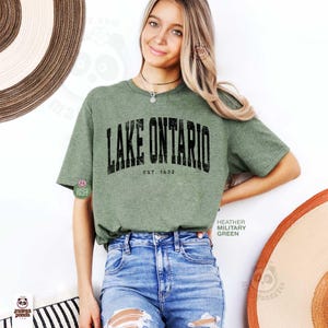 May include: A heather military green short-sleeve t-shirt featuring the black text LAKE ONTARIO and EST. 1632 printed in a distressed font across the chest. The shirt has a crew neckline and a standard fit, paired with blue denim jeans.