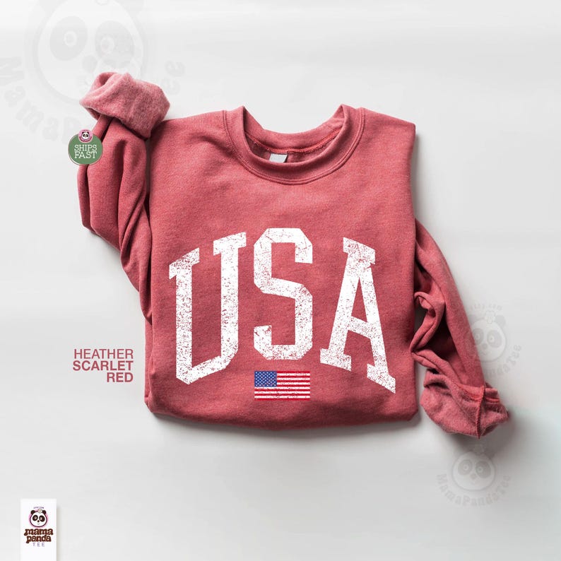 May include: Heather scarlet red sweatshirt with the word "USA" in distressed white lettering and a small American flag graphic. The sweatshirt has a crew neck and long sleeves.