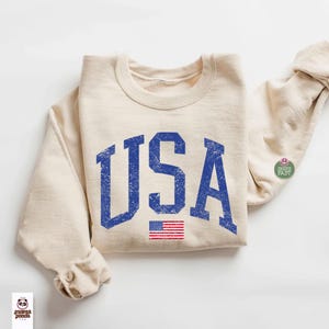 May include: Cream-colored sweatshirt with the word "USA" in distressed blue lettering and a small American flag graphic. The sweatshirt has a crew neck and long sleeves. A small "Ships Fast" sticker is on the sleeve.