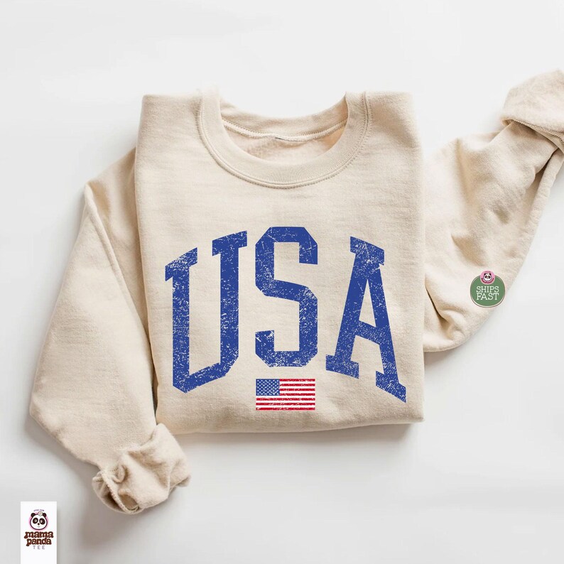 May include: Cream-colored sweatshirt with the word "USA" in distressed blue lettering and a small American flag graphic. The sweatshirt has a crew neck and long sleeves. A small "Ships Fast" sticker is on the sleeve.