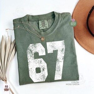May include: Moss green t-shirt with the number 67 in white, a gold necklace, and a brown hat. The shirt is labeled "Comfort Colors" and is size S-P-CH. The color is "Moss Green".