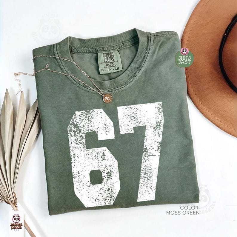 May include: Moss green t-shirt with the number 67 in white, a gold necklace, and a brown hat. The shirt is labeled "Comfort Colors" and is size S-P-CH. The color is "Moss Green".