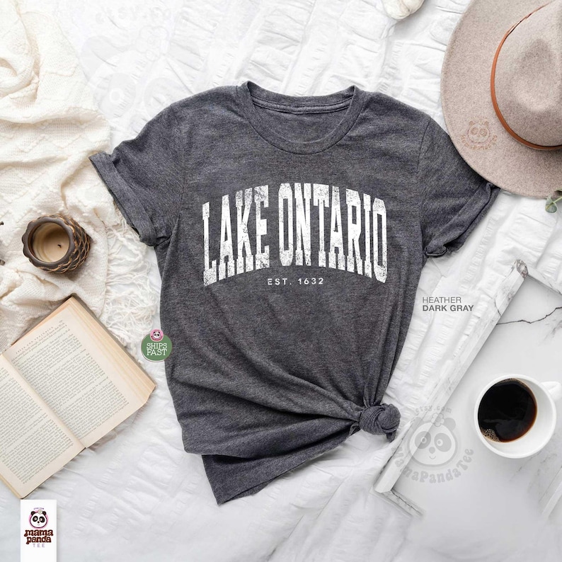 May include: A dark heather gray short-sleeve t-shirt featuring the white distressed text LAKE ONTARIO and EST. 1632 printed in an arched design across the chest. The shirt has a crew neckline and is made of a soft, textured fabric blend.
