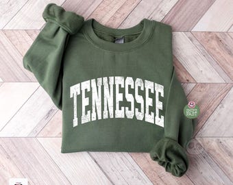 Op de afbeelding: Een olijfgroene sweatshirt met het woord "TENNESSEE" in witte, gebogen blokletters. De sweatshirt is op een licht houtnerfoppervlak gevouwen. De mouwen zijn opgerold. De afbeelding bevat een kleine "SHIPS FAST" sticker.