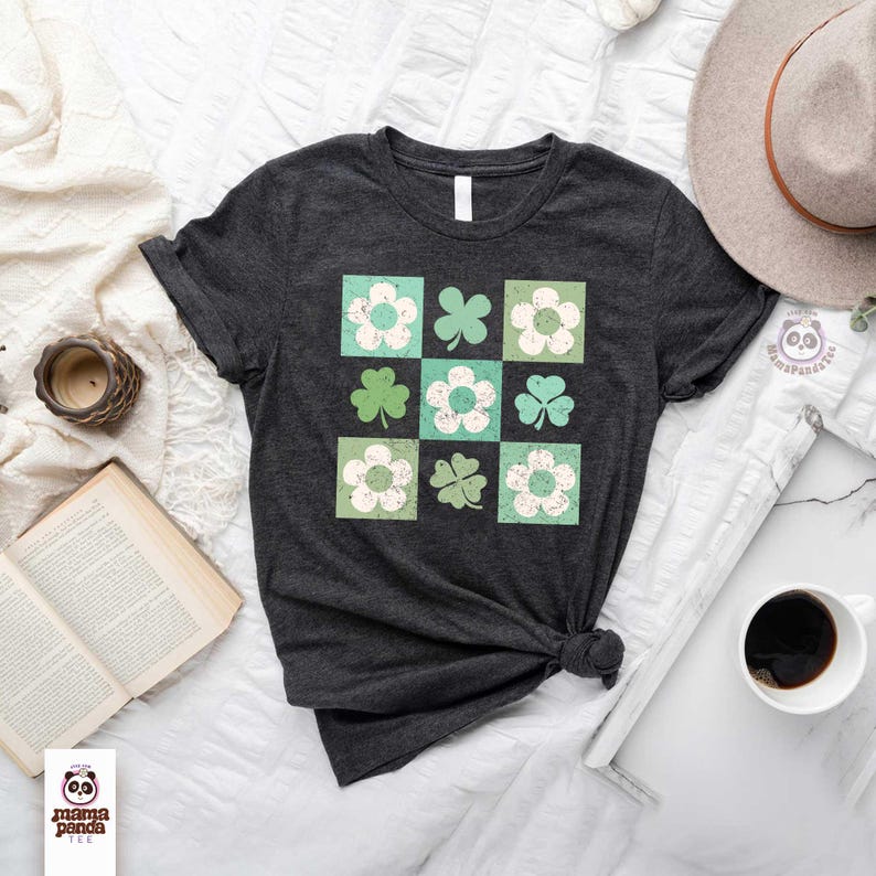 Op de afbeelding: Een donkergrijs T-shirt met een ontwerp van groene klavers en witte bloemen in een rasterpatroon. Het ontwerp heeft een vintage, verweerde look.