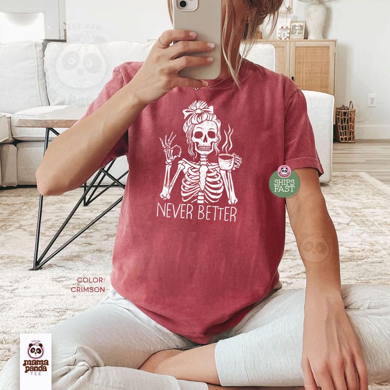 May include: A crimson short-sleeve t-shirt featuring a white graphic of a skeleton with a messy bun holding a coffee mug and making an okay hand gesture. The text "NEVER BETTER" is printed in white below the skeleton graphic on the front.