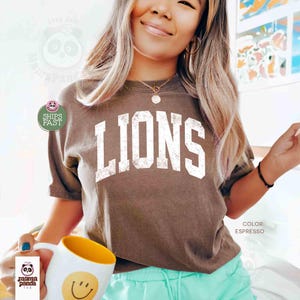May include: Espresso-colored t-shirt with the word "LIONS" in large, distressed white letters. The shirt is short-sleeved and has a crew neck. A white mug with a yellow interior and a smiley face is held in the foreground.
