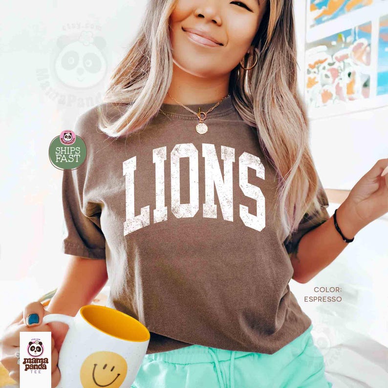 May include: Espresso-colored t-shirt with the word "LIONS" in large, distressed white letters. The shirt is short-sleeved and has a crew neck. A white mug with a yellow interior and a smiley face is held in the foreground.