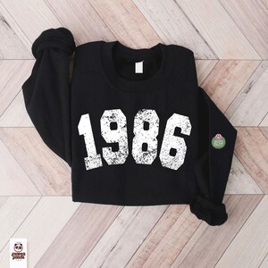 May include: Black sweatshirt with the year "1986" printed in distressed white lettering. The long-sleeved top has a crew neck and a small green and pink patch on the sleeve. The sweatshirt is laid on a light wood grain surface.