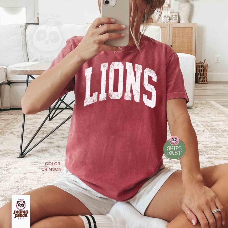 May include: Crimson-colored t-shirt with the word "LIONS" in distressed white lettering. The shirt is a crew neck style and appears to be made of a soft material. The text "COLOR: CRIMSON" is printed on the shirt. The shirt is part of the Mama Panda Tee collection.