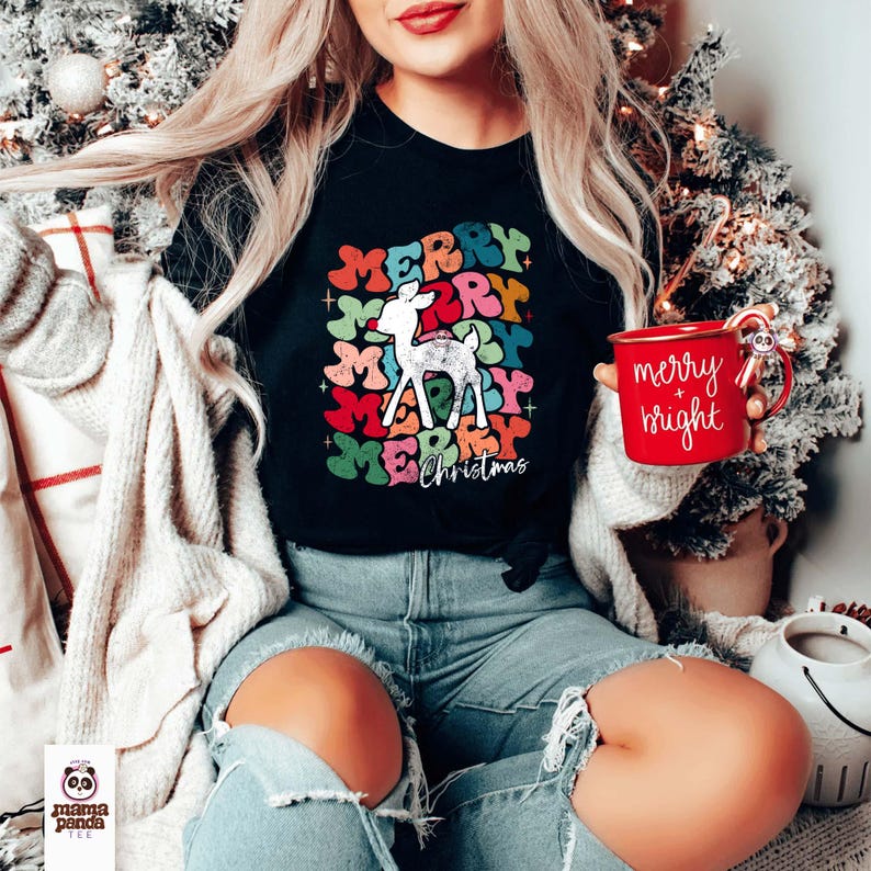 May include: A black t-shirt with a colorful "Merry Christmas" graphic featuring a white reindeer. The graphic has a vintage, distressed look. The shirt is being worn by a person holding a red mug with the text "merry + bright" in white.