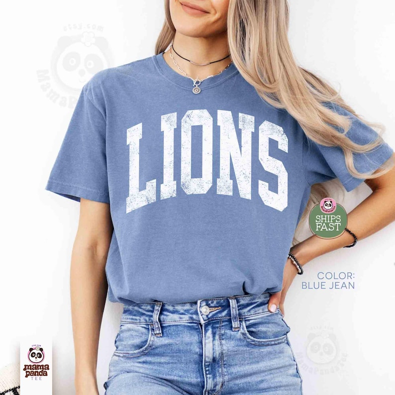 May include: Blue jean colored t-shirt with the word "LIONS" in distressed white lettering. The shirt has short sleeves and a crew neck. The text "SHIPS FAST" is visible on the right side of the shirt.